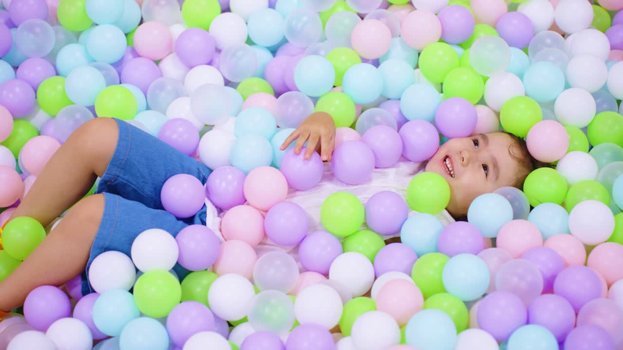 A joyful little girl lies in a colorful ball pit, laughing with excitement as a shower of pastel-colored plastic balls falls on her in slow motion at an indoor playground