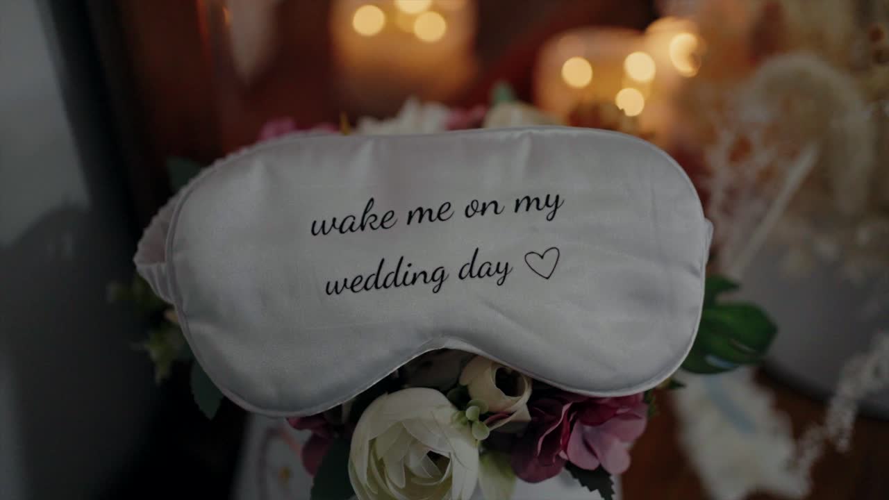 White sleep mask with "wake me on my wedding day" text, placed on a floral arrangement with soft lighting in the background