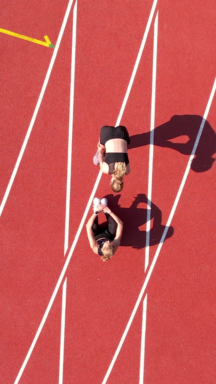 Toma vertical cenital de dos mujeres jóvenes con ropa deportiva estirándose y preparándose para correr en una pista de atletismo roja. Perfecta para contenido de fitness y redes sociales.