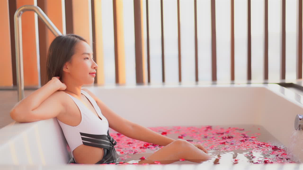 Pretty Asian model in black and white bathing suit soaking in spa tub with flower petals floating on surface. Woman relaxing in bath looking toward sunlight. Calming luxury resort amenities