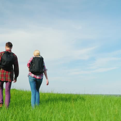 A-Couple-Of-Tourists-With-Backpacks-Are-Walking-Along-The-Crest-Of-A-Large-Green-Hill-2