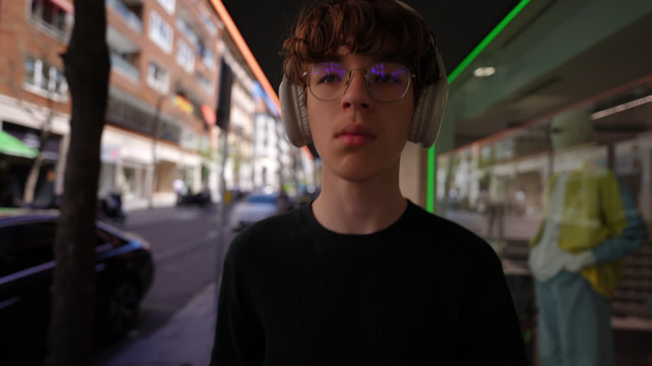 Teenager with headphones and glasses walks down a city street, looking straight ahead, seemingly lost in his thoughts as he enjoys his music