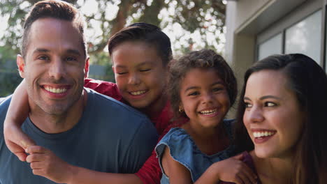 Portrait-Of-Smiling-Hispanic-Family-With-Parents-Giving-Children-Piggyback-Rides-In-Garden-At-Home