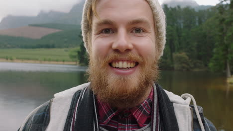 portrait red head hipster man with beard smiling feeling happy outdoors in nature by lake wearing beanie hat