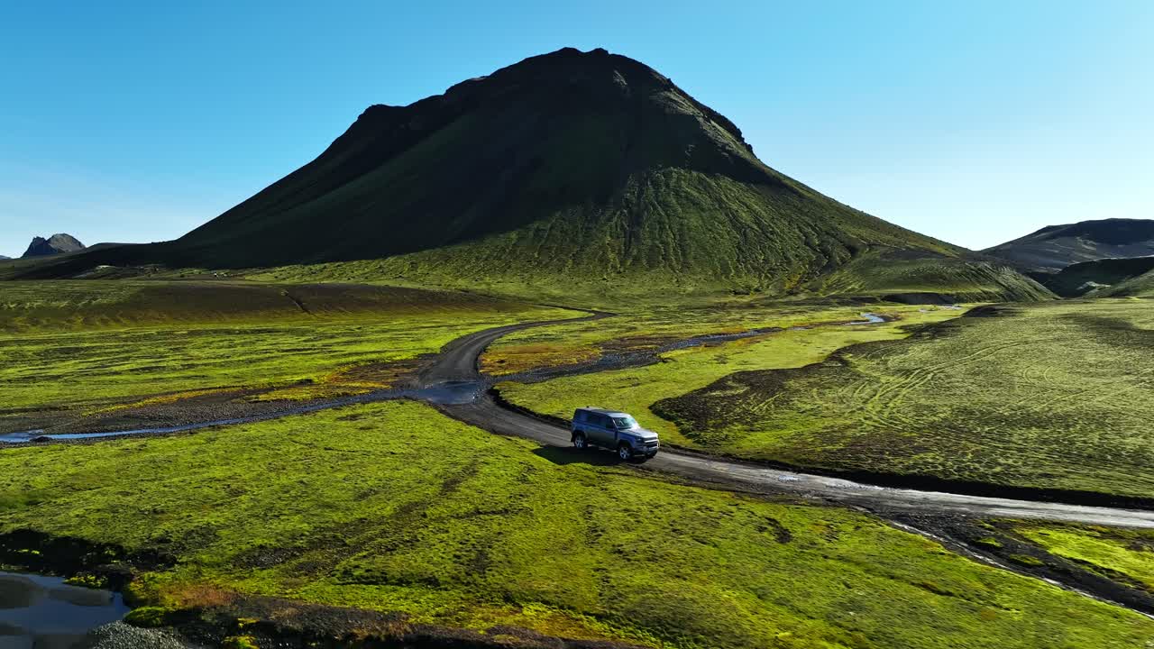 Vista drone di un SUV 4x4 che attraversa un guado poco profondo su una pista di ghiaia negli altopiani islandesi, con una ripida montagna verde e pianure vulcaniche sotto un cielo blu chiaro.