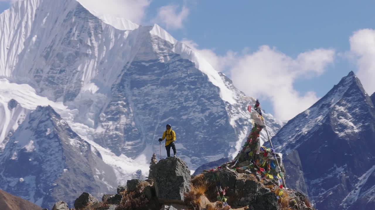 Drone shot with telephoto zoom of a male hiker reaching the summit in Nepal Mountain. Surrounded by high-altitude snow-capped Himalayas including Everest and Langtang, adorned with prayer flags.