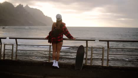 Full-Length-Sporty-Builded-Woman-In-Hat,-Plaid-Coat-And-White-Sneakers-Enjoying-Time-By-The-Seaside-On-A-Cloudy-Day-At-Sunset,-Standing-Leaning-On-Railings-With-Skateboard,-Watching-Stormy-Ocean-From-Promenade