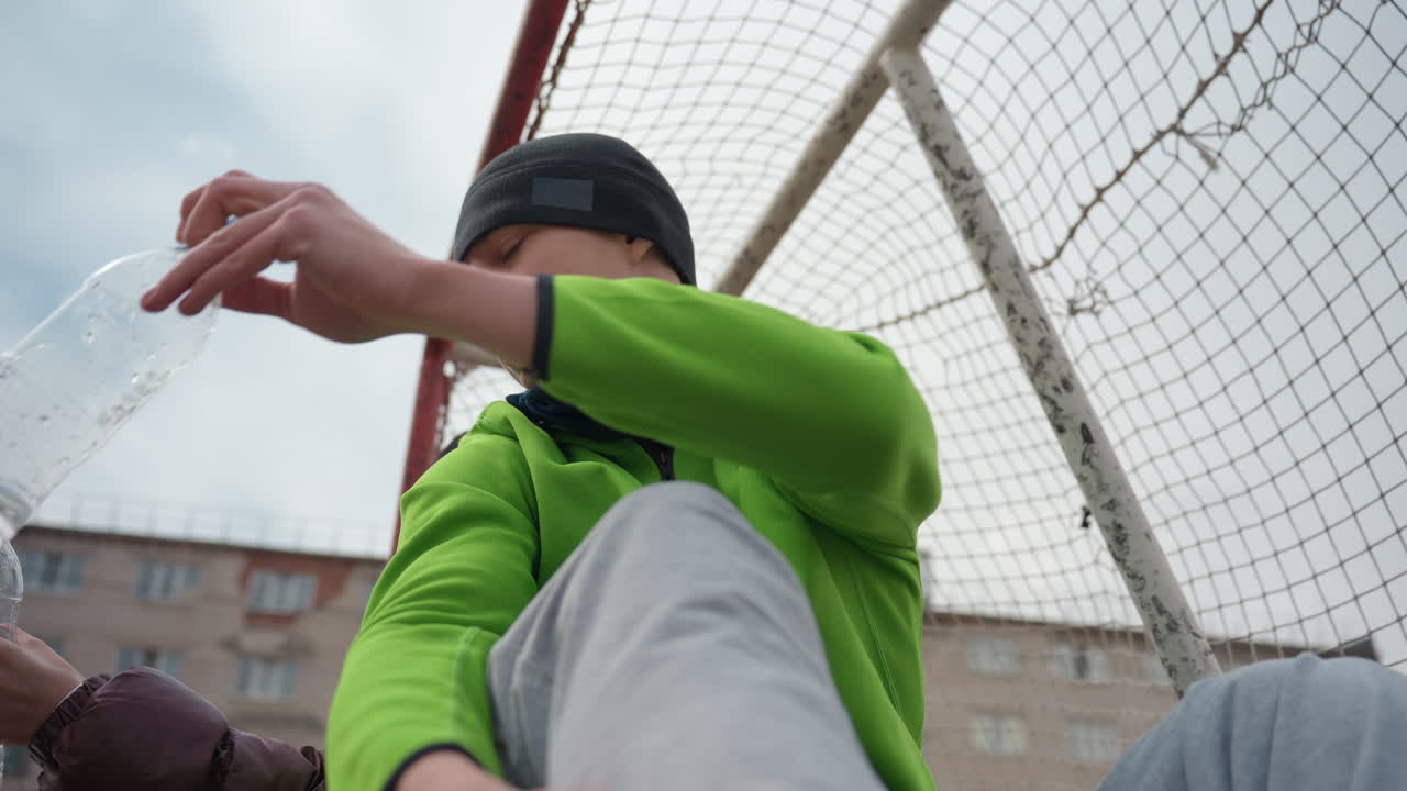 ゴールポストの近くでボトルを開ける男性。ゴールポストのそばで休憩しながら水筒を扱う人。練習休憩中にウォーターボトルのキャップを開け、スポーツ用ゴールのそばでくつろぐ白人男性。
