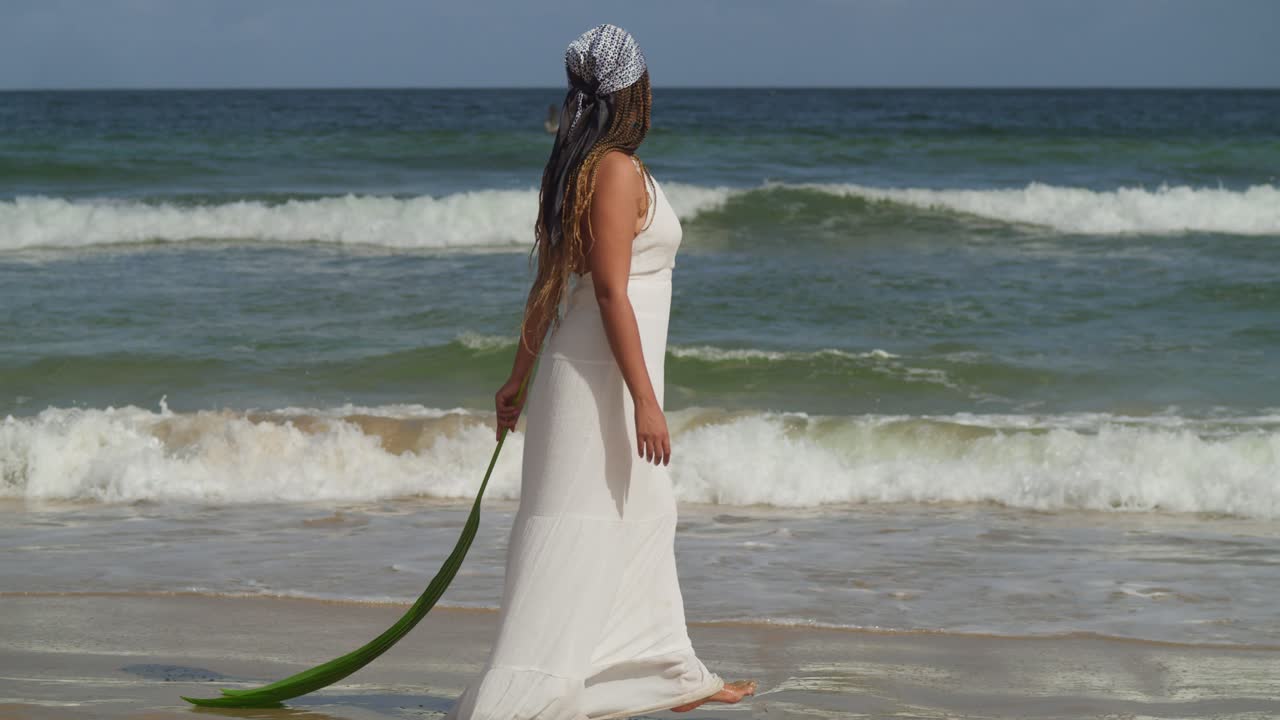 niña caminando en la playa sosteniendo una hoja de palma mientras usa un vestido de playa y olas del océano rompiendo la costa en el fondo