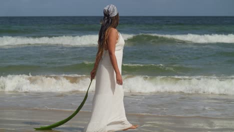 jong meisje dat op het strand loopt en een palmblad vasthoudt terwijl ze een strandjurk draagt en oceaangolven die op de achtergrond de kustlijn beuken