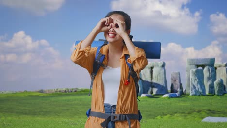 asian female hiker with mountaineering backpack making binoculars gesture looking at something then saying wow while traveling in stonehenge
