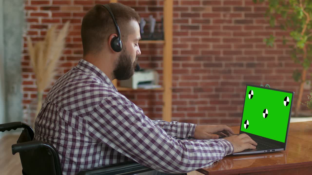 hombre en silla de ruedas con auriculares está sentado en la computadora portátil con chromakey y trabajando, vista trasera. persona con discapacidad trabaja remotamente. oportunidades de concepto