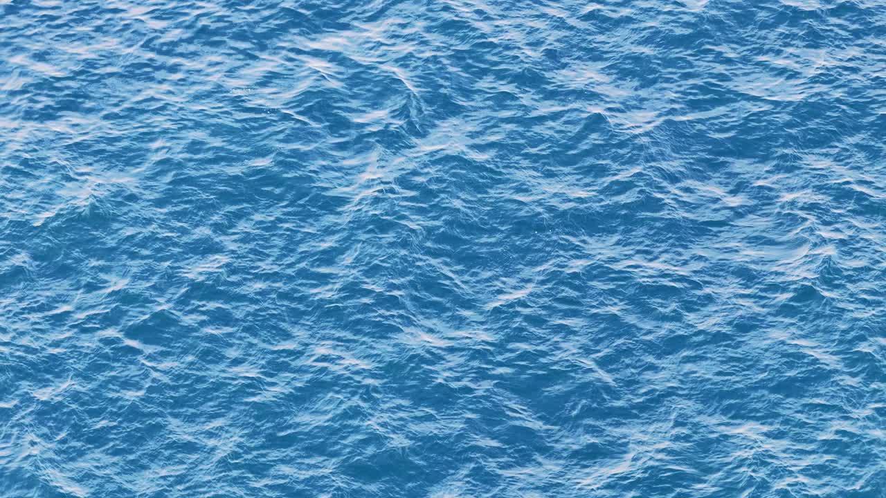 High-resolution aerial footage captures dynamic, sunlit ocean surface with rough blue waves. Smooth drone movement creates a sense of vastness and natural energy