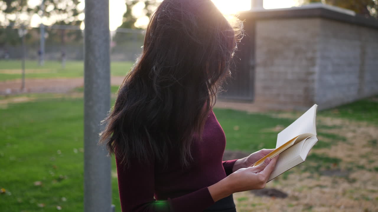 una joven estudiante universitaria leyendo un libro de texto al aire libre en el parque del campus al atardecer a cámara lenta