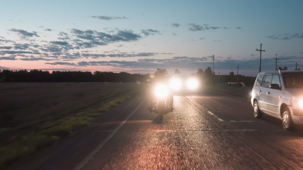 Security agent on motorcycle traveling rural road during dusk, cars following from behind with bright headlights, one vehicle overtaking while trees and open field frame background