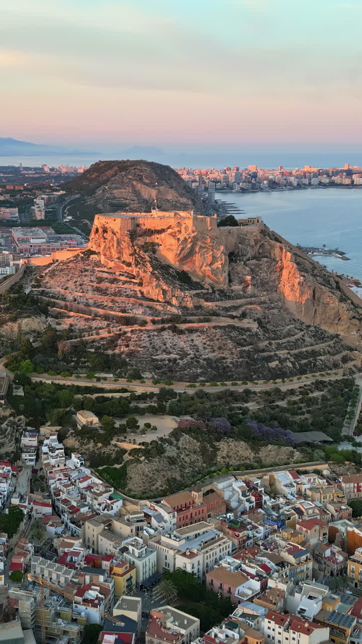 Dronebillede i fugleperspektiv af Santa Barbara-slottet omgivet af bygninger ved kysten af Alicante, Spanien, med havet i baggrunden. Lodret