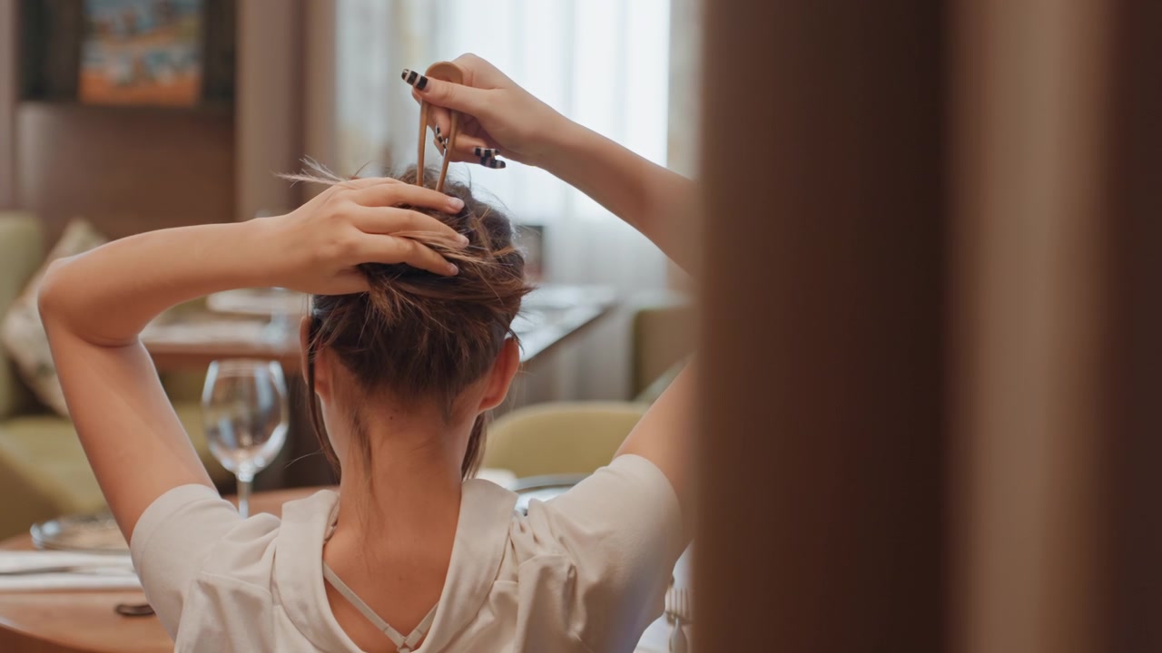 Mujer peinándose el cabello en un moño durante la preparación nocturna, con un comedor íntimo de fondo y luz ambiental suave, practicando una técnica de recogido con horquillas y pasadores, manos concentradas y un ritual seguro.