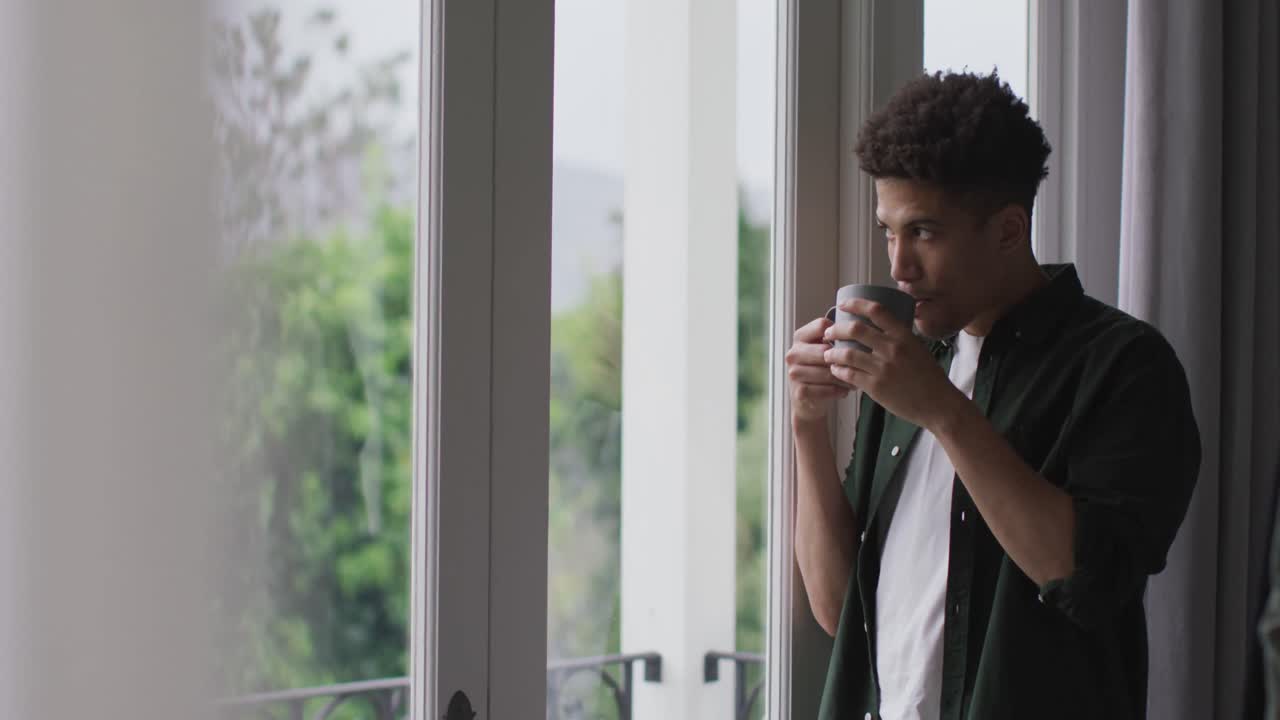 Uomo di etnia mista pensieroso che guarda fuori dalla finestra e beve caffè.