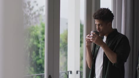 thoughtful biracial man looking through the window and drinking coffee