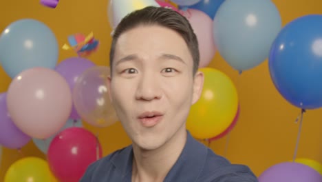 pov studio portrait of man taking selfie wearing party hat celebrating birthday surrounded by balloons