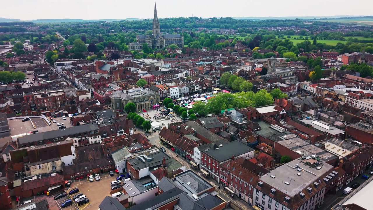 Drone footage looking across Salisbury city centre before a public civic event with Market Square rooftops historic buildings greenery and Salisbury Cathedral in the distance.