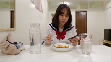 Portrait-Of-A-Young-Girl-Wearing-Uniform-Is-Eating-Dinner-While-Using-Smartphone