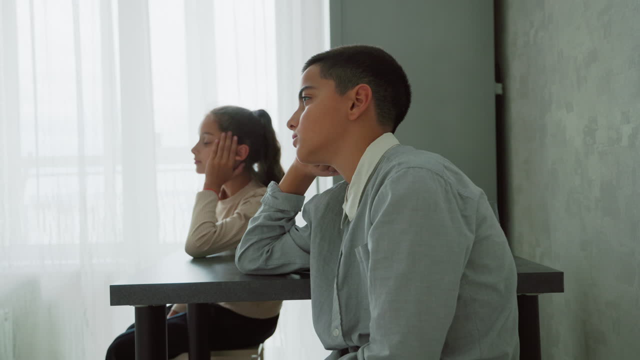 children in school uniforms sitting at table near sheer curtain window in morning light looking thoughtful and calm while waiting indoors during quiet home routine before heading to school