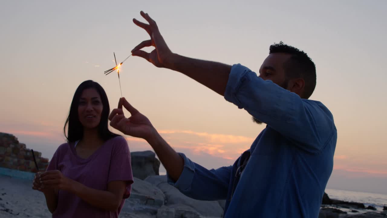 joven pareja feliz de raza mixta divirtiéndose con chispas en la playa 4k