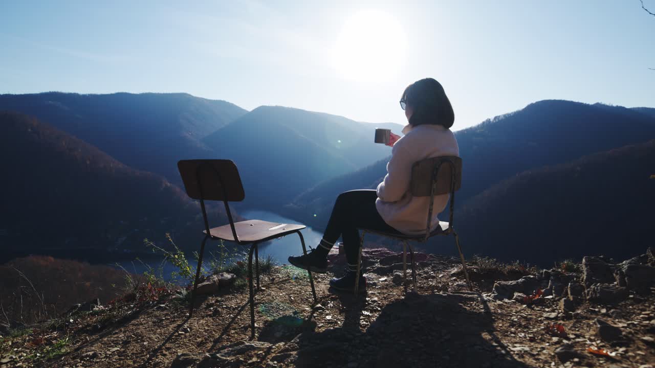 A static, wide shot of a woman sitting on a metal chair on a cliff edge, drinking from a mug and looking over a vast mountain valley and river. Conveys solitude and freedom