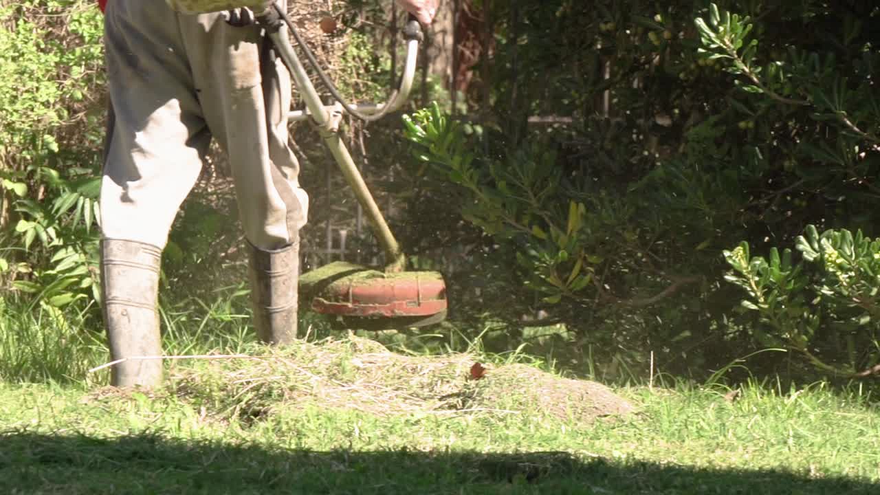 Um homem trabalha em um jardim ensolarado cortando grama alta com um cortador de mato durante o verão. Câmera lenta