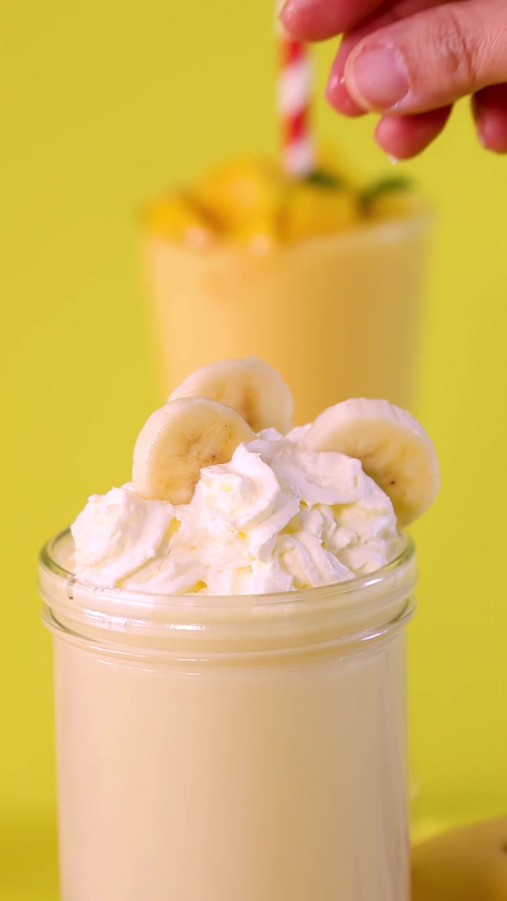 Hand places banana slices on whipped cream-topped banana milkshake, bright yellow background, close-up