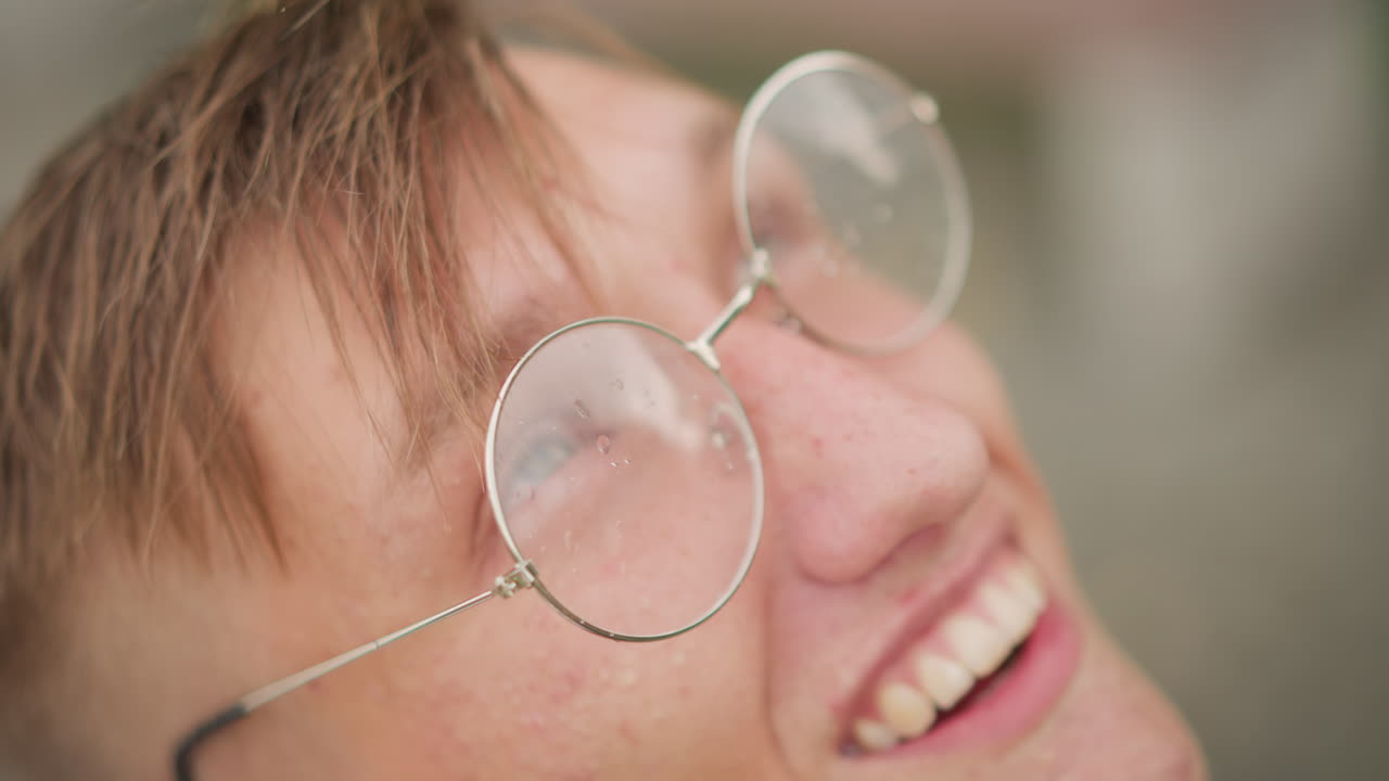 primer plano de un joven caucásico con gafas riendo, retrato alegre con lentes redondas que magnifican las gotas de lluvia, sonrisa espontánea, escena urbana al aire libre, energía despreocupada y ambiente de amistad bajo una ligera llovizna