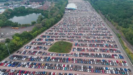 a large car park with neatly arranged cars in various colours, surrounded by greenery and a waterbody, with an urban area in the background