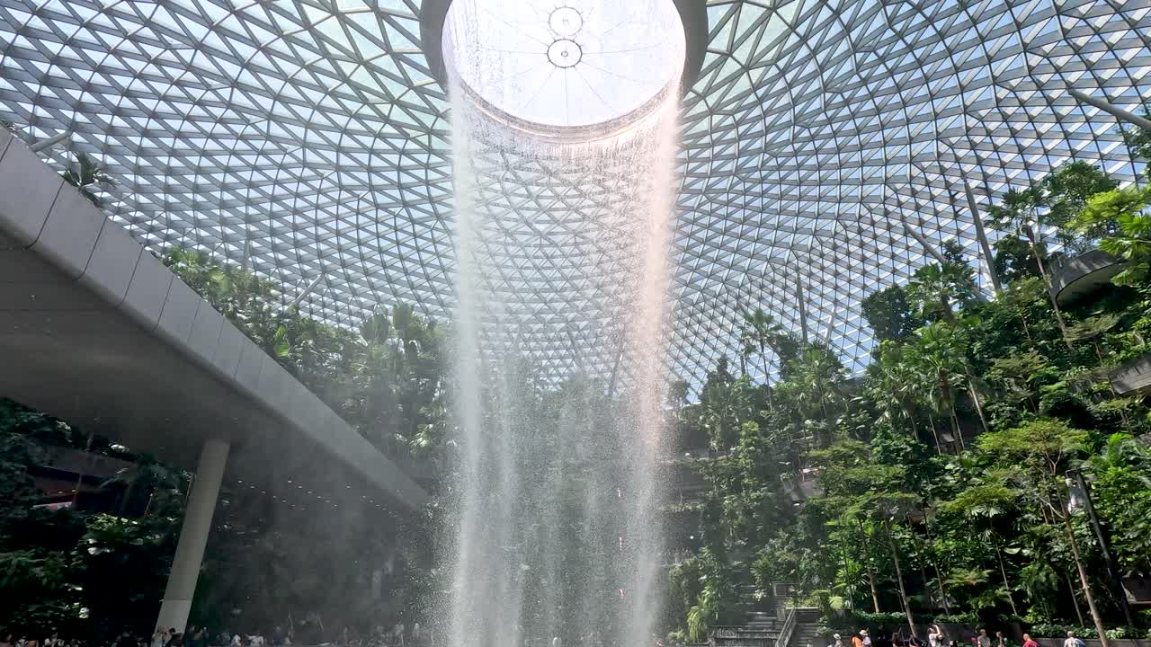 Tall indoor waterfall flows beneath geometric glass dome, surrounded by lush greenery and natural daylight