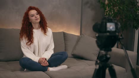 Caucasian-red-headed-young-girl-vlogger-is-talking-in-front-of-camera-recording-video-for-online-blog-for-her-followers,-smiling-and-gesturing.-Woman-is-wearing-jeans-and-white-sweater.-Accelerated-video.-Camera-on-foreground