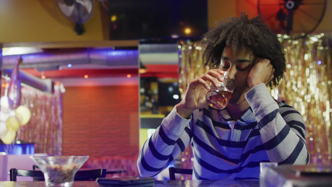 Video of drunk african american man sitting drinking at the bar and bowing head down
