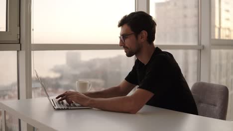 joyful man using laptop computer at remote workplace in slow motion. freelancer working on computer at home. good looking man actively typing on laptop, chatting with friends at big house with panoramic windows. close up