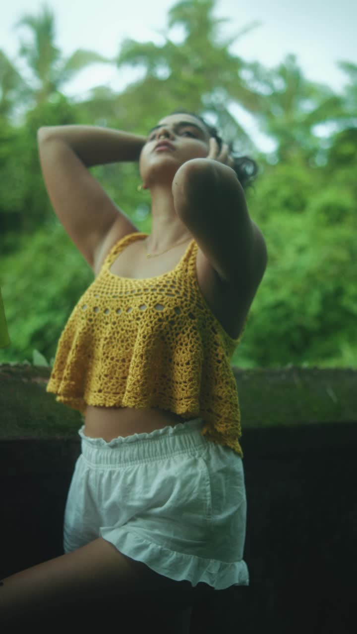 Vertical shot of woman in yellow crochet top and white shorts, raising hair and posing sensually outdoors