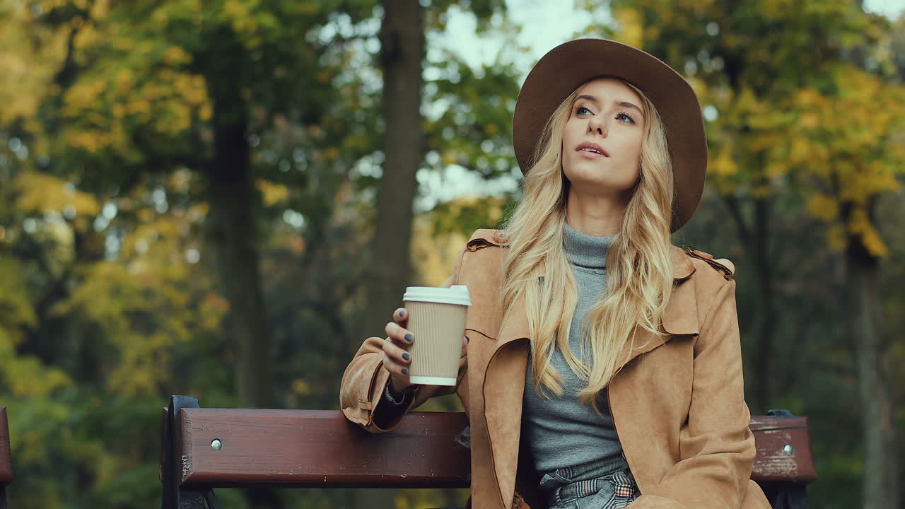 Caucasian blonde caucasian woman sitting on the bench in a park, drinking coffee while waiting for somebody and looking at the watch