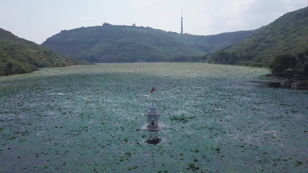 Luchtfoto van het Jait Sagar Meer in Bundi, met een klein tempelpaviljoen met rode vlag te midden van uitgestrekte lotusbladbedekking, ingekaderd door groene heuvels en een verre toren onder een bewolkte hemel