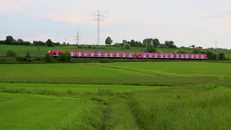 Red-train-crossing-lush-green-countryside-with-power-lines-in-background,-daytime