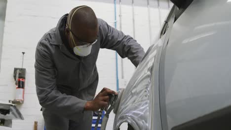 Afroamerikanischer-Männlicher-Automechaniker,-Der-Eine-Gesichtsmaske-Trägt-Und-Eine-Seite-Eines-Autos-Mit-Einer-Schleifmaschine-Poliert