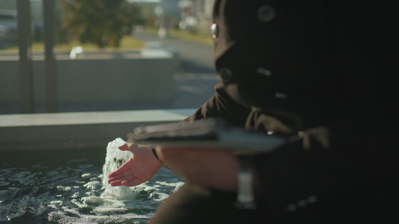 검은 코트를 입은 남자가 야외에 앉아서 밝은 빛 아래 물 근처의 태블릿과 함께 다중 작업을 하고 있습니다. 부드러운 보케 배경으로 활발한 몸짓으로 손을 고 있습니다.