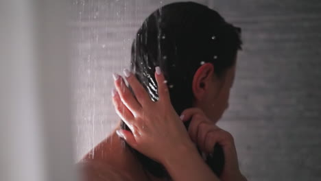 woman washes away shampoo from hair shower unit. brunette lady strokes short hair under falling down water drops in bathroom. regular procedure