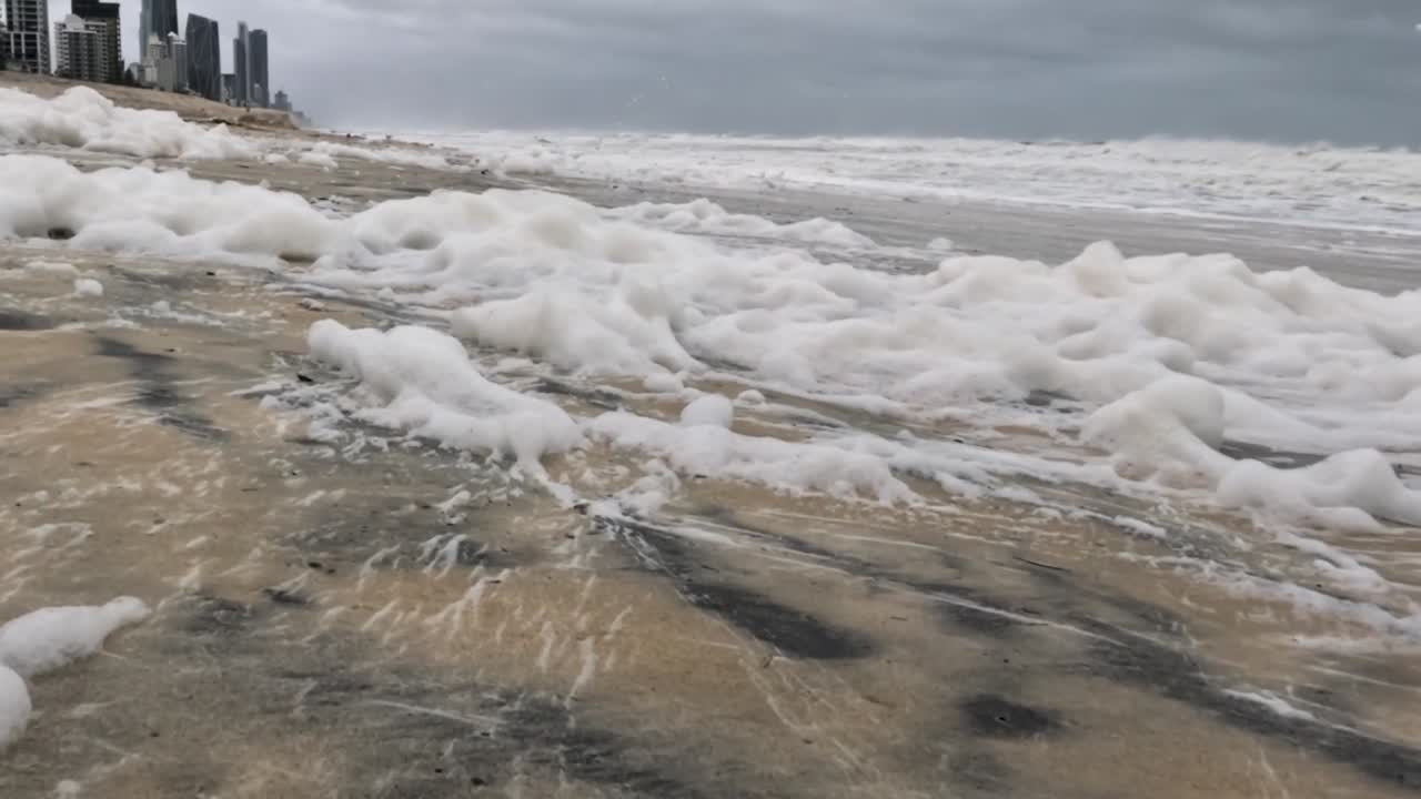 Close-up view of sea foam on a sandy beach with distant city buildings under cloudy skies.
