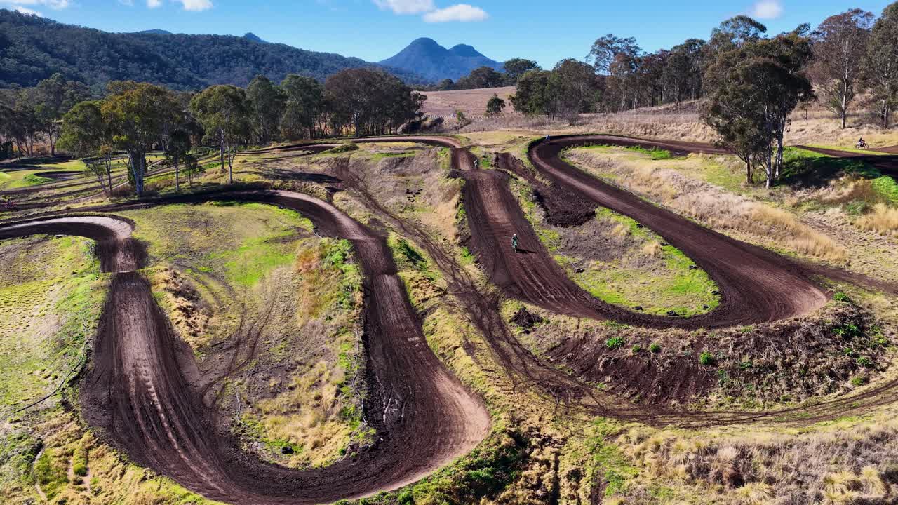 Motorbikes race on winding dirt track through bushland scenery