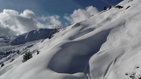 Drone-shot,-pedestal-up-from-behind-snow-humps-to-reveal-a-snow-covered-mountain-side-with-rocks-and-sunny-sky-in-La-Plagne-,-French-Alps