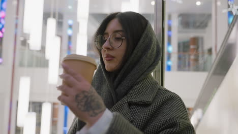 young woman in glasses and houndstooth coat stands in glass elevator sipping juice, surrounded by bright hanging lights and modern decor as elevator ascends through stylish shopping mall interior