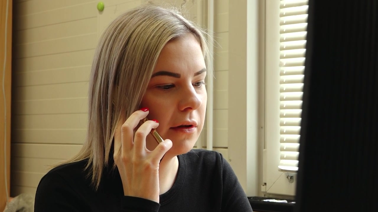 Woman sits at a desk using a computer while talking on a smartphone in a small home office with patterned walls and focused work atmosphere.