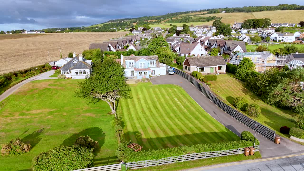 Drone camera glides above Rosemarkie, revealing residential homes, manicured lawns, and golden farmland under bright daylight with scattered clouds